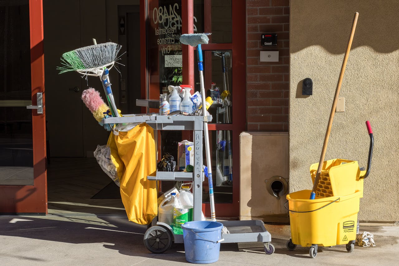 about-img-01 Cleaning supplies and tools outside a building entrance bathed in sunlight.